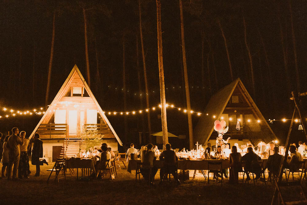 Slow wedding w lesie, goście siedzą przy stołach. W tle widoczne chatki.